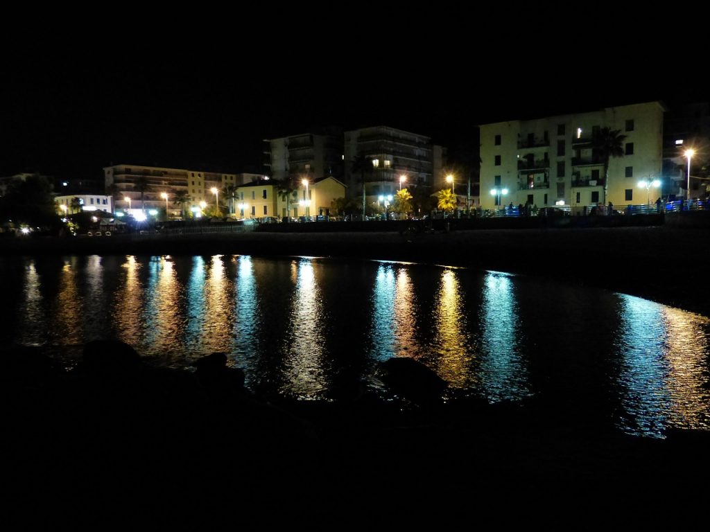 A photo of a port city's buildings. Light coming from the streets is reflecting in the black water.